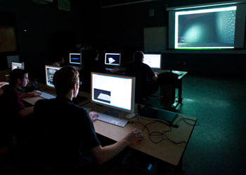 Students in dark room look at computer screens and big screen in front of room
