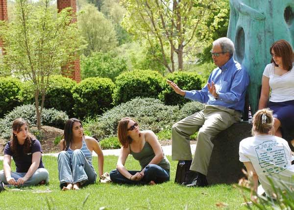 Roger Wilkins rests on the plinth of &quot;Communitas&quot; environmental sculpture on the Fairfax campus of 番茄视频. He is speaking to a group of students. It is a beautiful sunny spring day, and students listen attentively as they are seated on the ground. Wilkins is wearing khaki trousers, a blue oxford shirt, and sunglasses.
