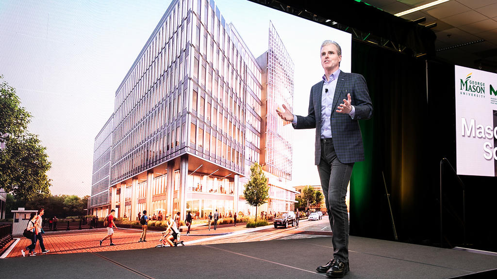 Paul Misener stands on a stage in front of a projected image at the Mason Square groundbreaking event. The image is concept art for the Fuse building.