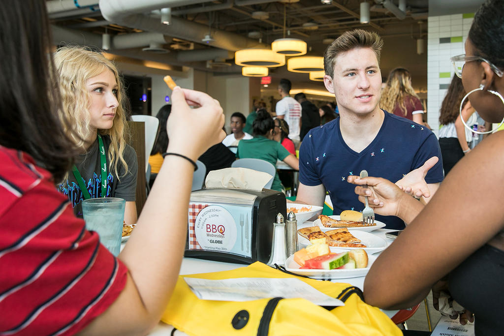 Four students sit around a table in a very busy dining hall. They are having an active conversation in the middle of their dining.