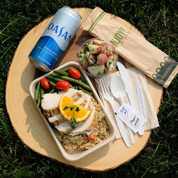 An example of a meal for those who are too sick to eat in a dining hall. The main container of food contains slices of grilled chicken, couscous, green beans, and cherry tomatoes, all in a compostable container. Beside this is a container of potato salad, a can of Dasani water, silverware, salt and pepper packets, and a napkin.