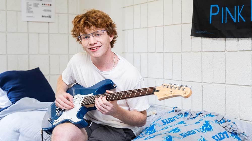 student playing guitar on his bed in dorm room.