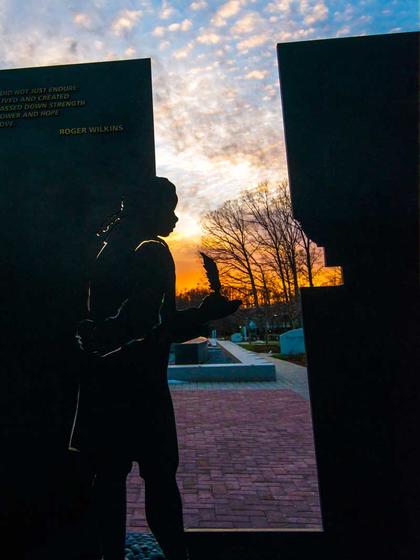 The memorial is constructed out of verticle steel plates, which are cut to reveal the silhouettes of people enslaved by George Mason. In this photo, Penny, a young girl is shown appearing to walk through a doorway, delivering writing supplies to Mason.